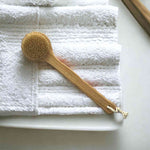 Eco-friendly body brush with natural bristles and a wooden handle, resting on white textured towels, perfect for skin exfoliation and lymphatic drainage.