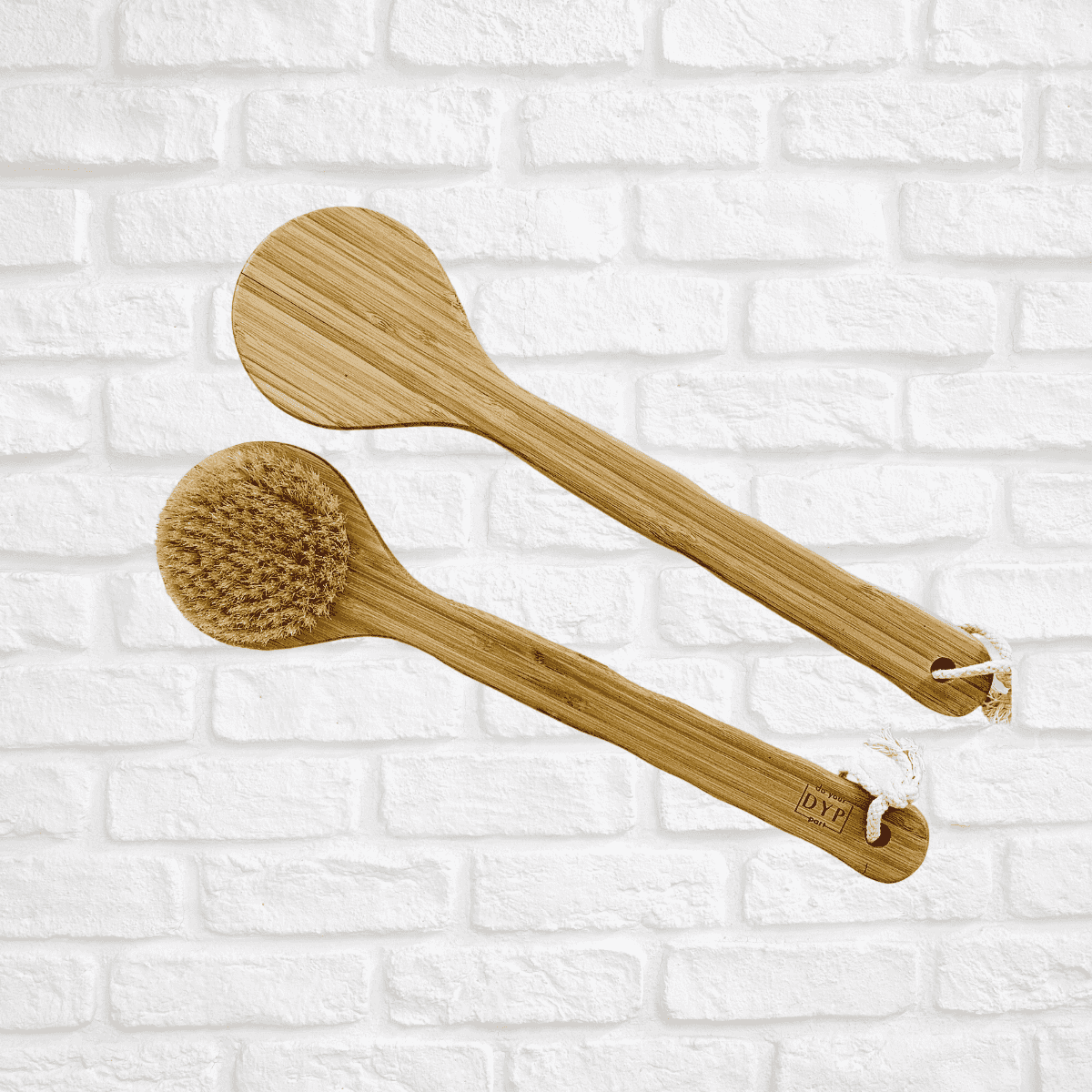 Set of two eco-friendly body brushes, one with natural bristles and one flat, both with wooden handles, displayed against a white brick background.